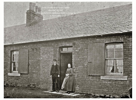 Photo:Archibald Dundas and his wife, great grandparents of Mrs Meg Stenhouse.  Standing outside their house at Muirhall, before 1911.  Archie worked with the Caledonian Railway Company in West Calder.
